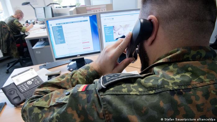 Ein uniformierter Bundeswehr-Soldat telefoniert sitzend an einem Schreibtisch mit zwei Computer-Bildschirmen. 