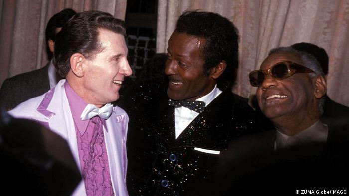 Jerry Lee Lewis, Chuck Berry und Ray Charles stehen in Anzügen nebeneinander und lachen gemeinsam.