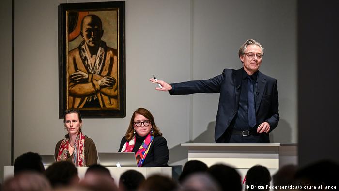 Ein Auktionator zeigt auf Max Beckmanns Selbstbildnis gelb-rosa im Auktionshaus Grisebach, zu seiner rechten sitzen zwei Frauen am Laptop.