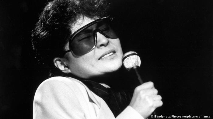 Yoko Ono mit Sonnenbrille, sie singt in ein Mikrofon.