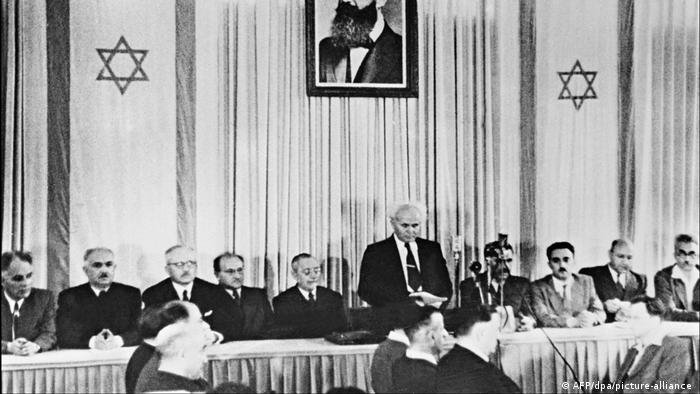 Auf einem Schwarz weiß Foto ist stehend David Ben Gurion zu sehen, neben ihm sitzen zahlreiche Männer, hinter ihm ein Portrait Theodor Herzl und die israelische Flagge